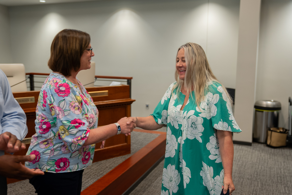 Two women shake hands in a conference room with a wooden desk. One woman wears a green floral dress and the other a pink floral blouse.