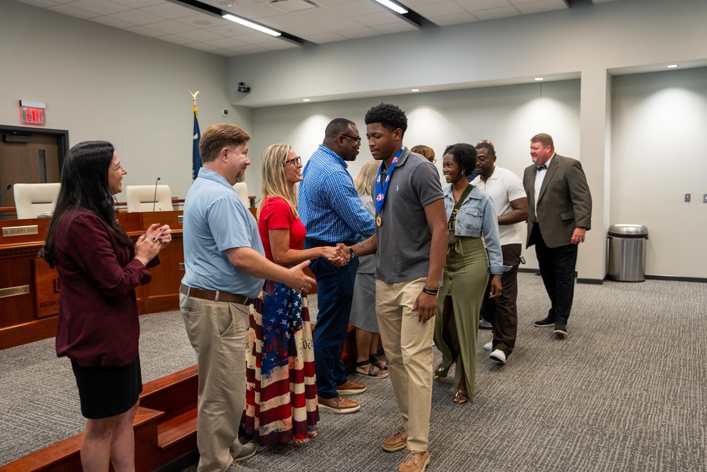 Group of people in a conference room, shaking hands. Some wear casual attire; others wear business suits.