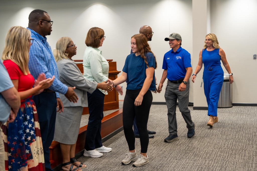 Group of adults, some in business attire, stand in a room with a raised platform. A woman in blue shakes hands with another woman.