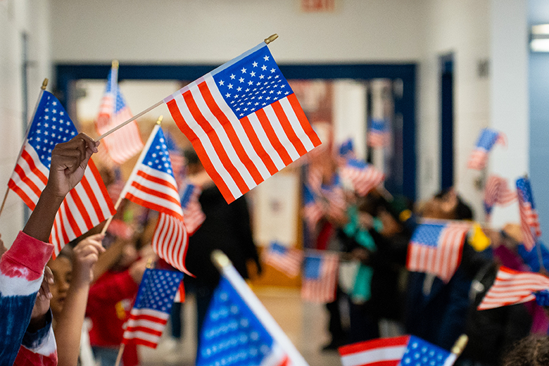 Children hold small American flags in a classroom setting. Some flags are blurry in the foreground and background.