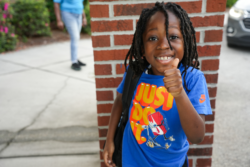 Child giving thumbs up stands next to brick wall. Another person walks on the sidewalk behind the child.