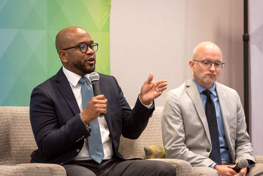 Two men in suits and ties, one holding a microphone, seated on a couch. One man gestures. Behind them, a multicolored banner.
