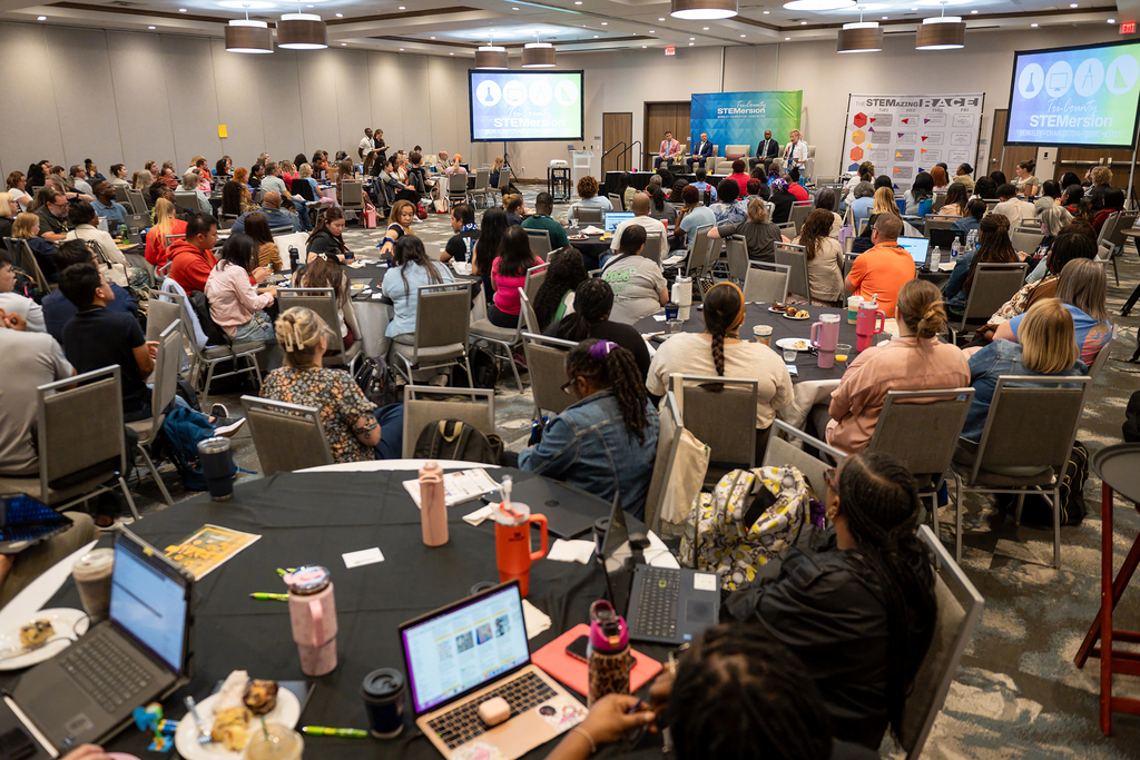 A room with a crowd of people seated at round tables. Laptops, cups, and plates are on the tables.