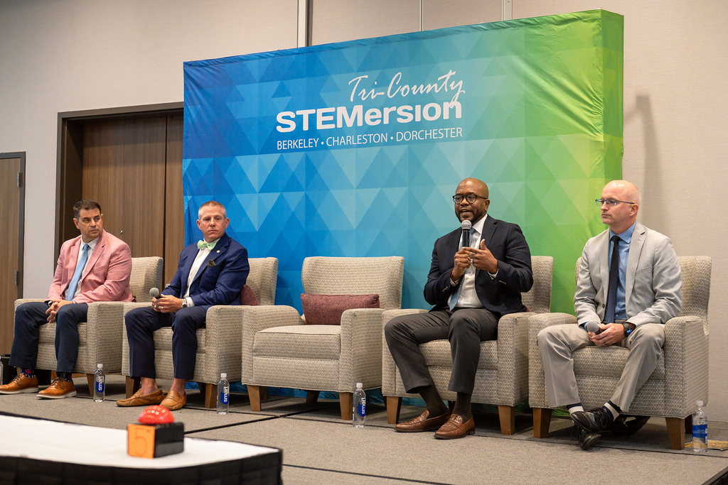 Four men in suits sit on chairs on a stage. One man holds a microphone. A banner reads "TenCounty STEMersion."