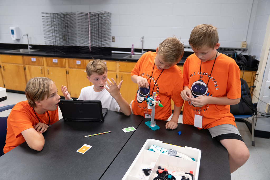 Four boys in orange shirts sit around a table. One looks at a laptop, another holds a robot model, and the other two watch.