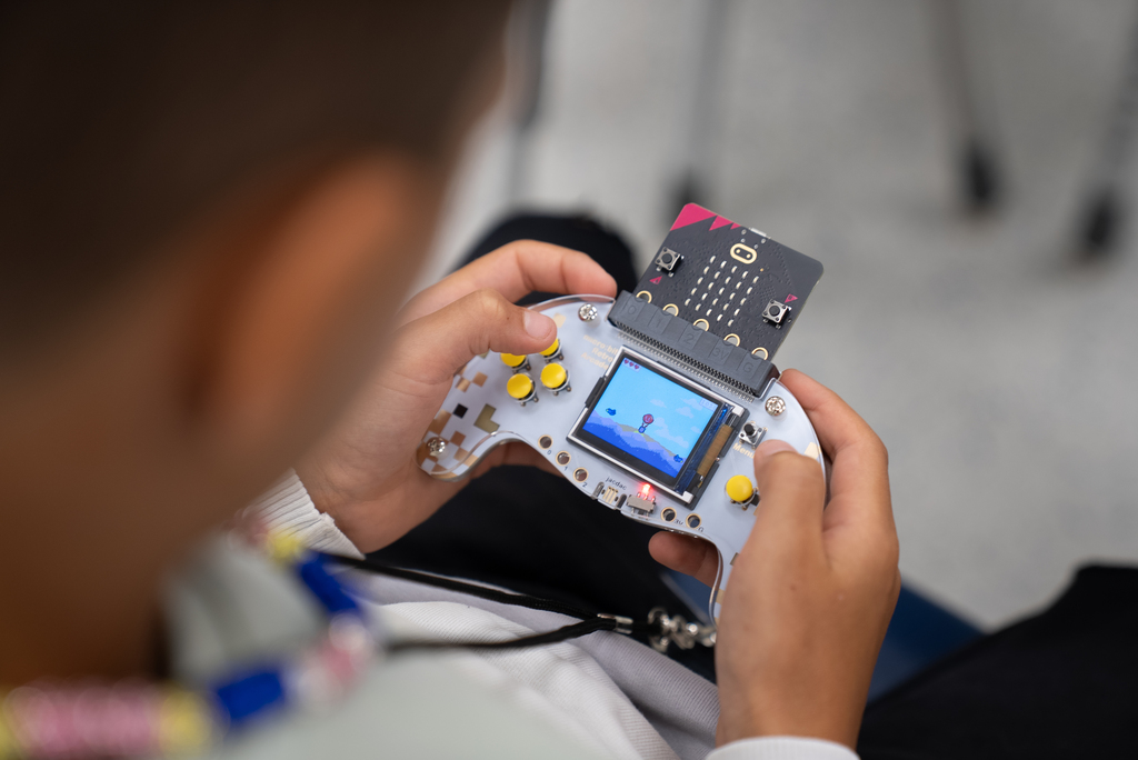 A person holds a small electronic device with a screen and buttons. They are seated in a room.