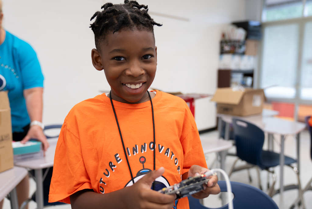 A smiling child in an orange shirt holds a game controller while another person stands behind a table.