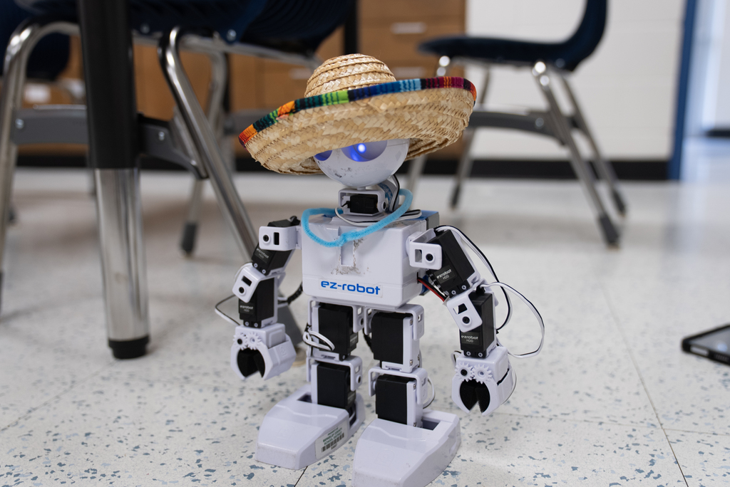A robot with a straw hat sits on a floor. Chairs and a table are in the background.