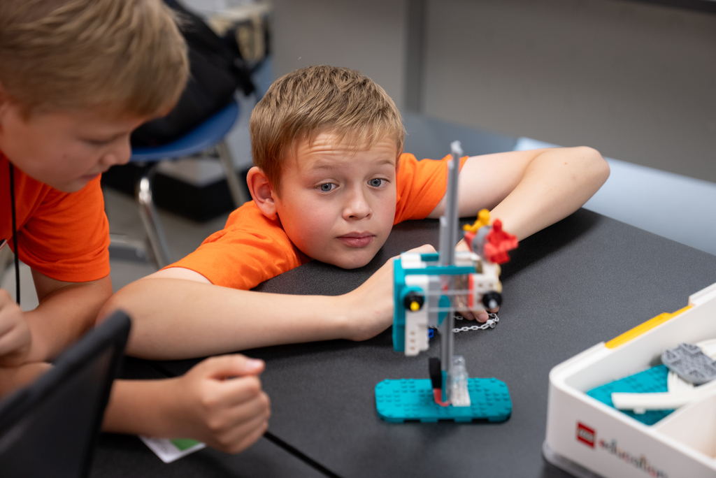 Two children, one with blond hair, are seated at a table, intently working with a Lego structure.