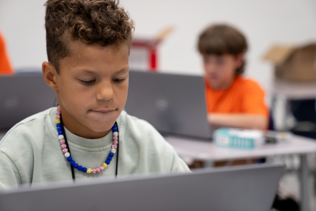 A boy in a gray top with a colorful beaded necklace sits at a desk, looking at a laptop.