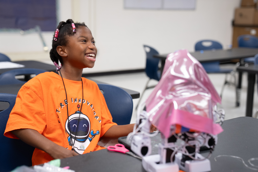 A child smiles, seated at a desk, working on a craft project with scissors and a robot model.