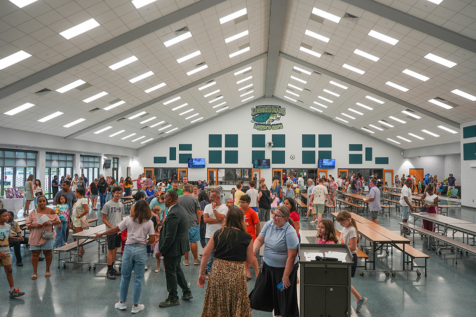 Large room with many people. Tables and chairs arranged. Bright lighting from ceiling. People moving around.