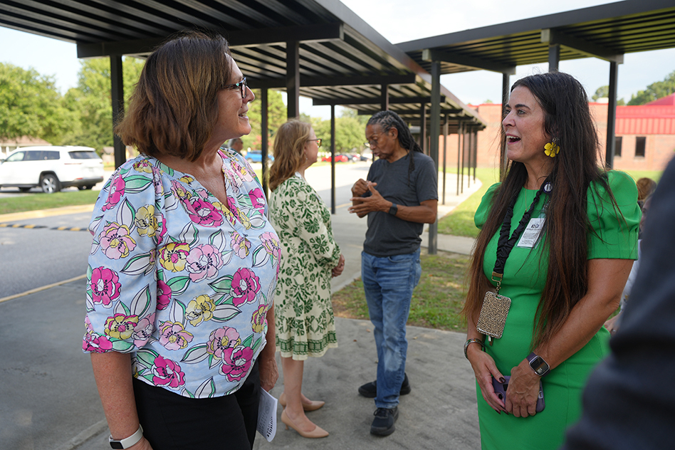 Four adults in a school parking lot. One wears a floral top, and another wears a green dress.
