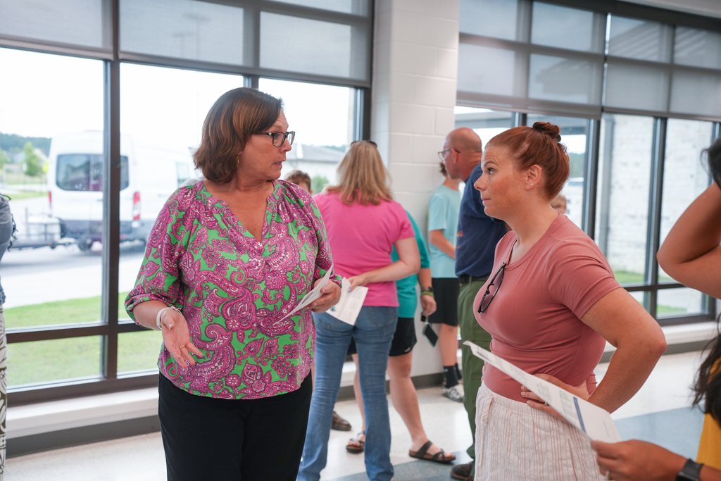 Several people stand in a room. One woman in floral top speaks to another woman with a ponytail.