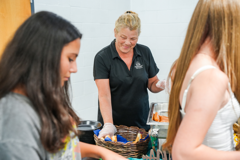 A woman in black serves food to two girls in gray and white, against a white wall.