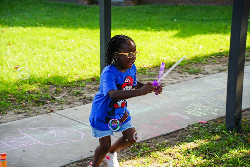 A child wearing a blue shirt and glasses holds two bubble wands while running on a sidewalk.