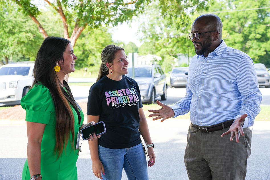 Three people outdoors in a parking lot. One man speaks while gesturing. Two women listen.