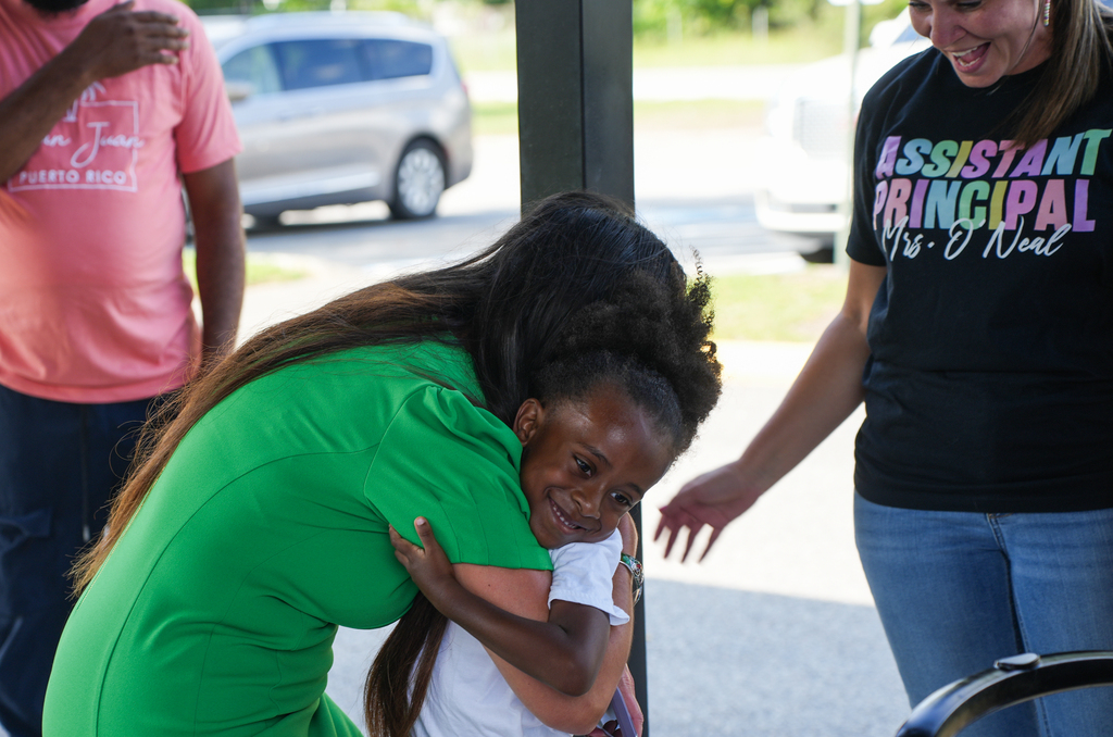 A woman hugs a girl in a green shirt, a woman in a black shirt holds her arm, and another person stands behind them.