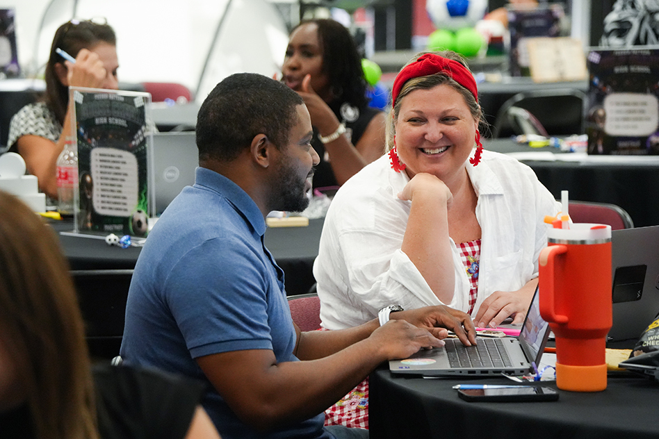 Two people are seated at a desk, working on laptops. One person wears a blue shirt, the other wears a white coat.
