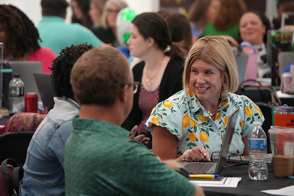 People at tables in a room, two wearing glasses, with laptops and water bottles, one woman smiling.
