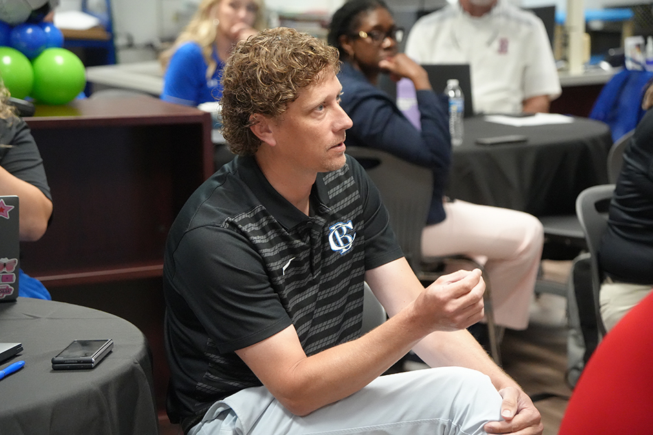Man with curly hair, seated in a meeting with a black and white striped polo.
