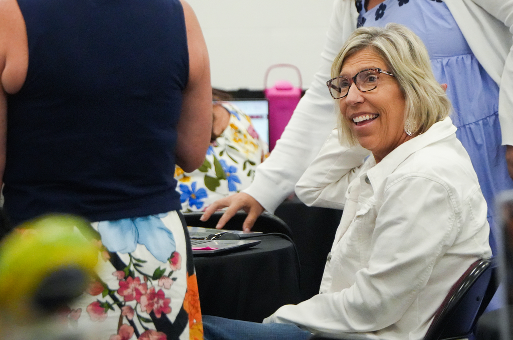 A woman in a white shirt sits in a chair smiling, next to another woman in a blue dress.