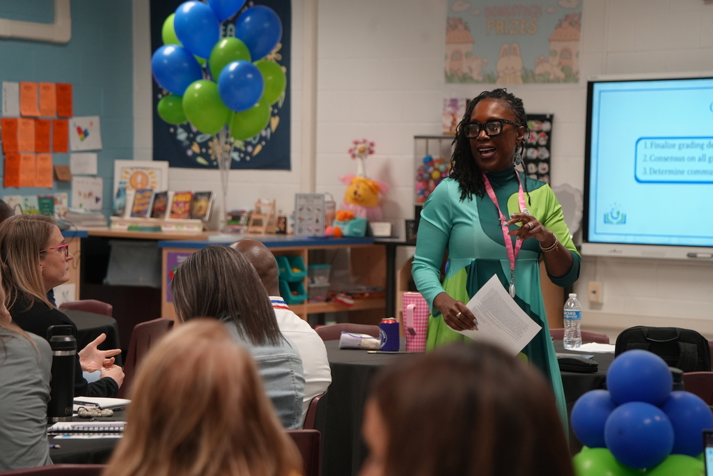 Woman in green dress presenting to seated group in classroom with balloons, a screen, and bulletin board.