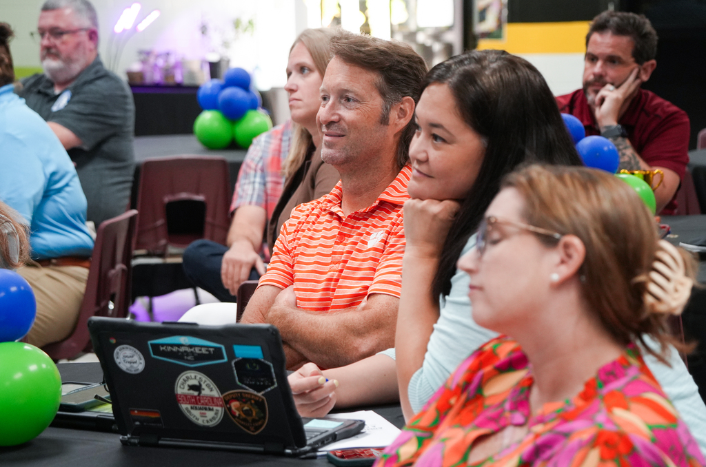 A group of adults sit around a table with a laptop. One man wears glasses, and others have their arms crossed.