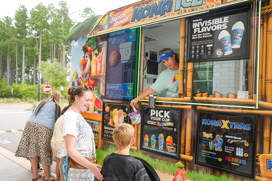 People standing outside a Kona Ice truck, a man behind the window, a menu board on the side.