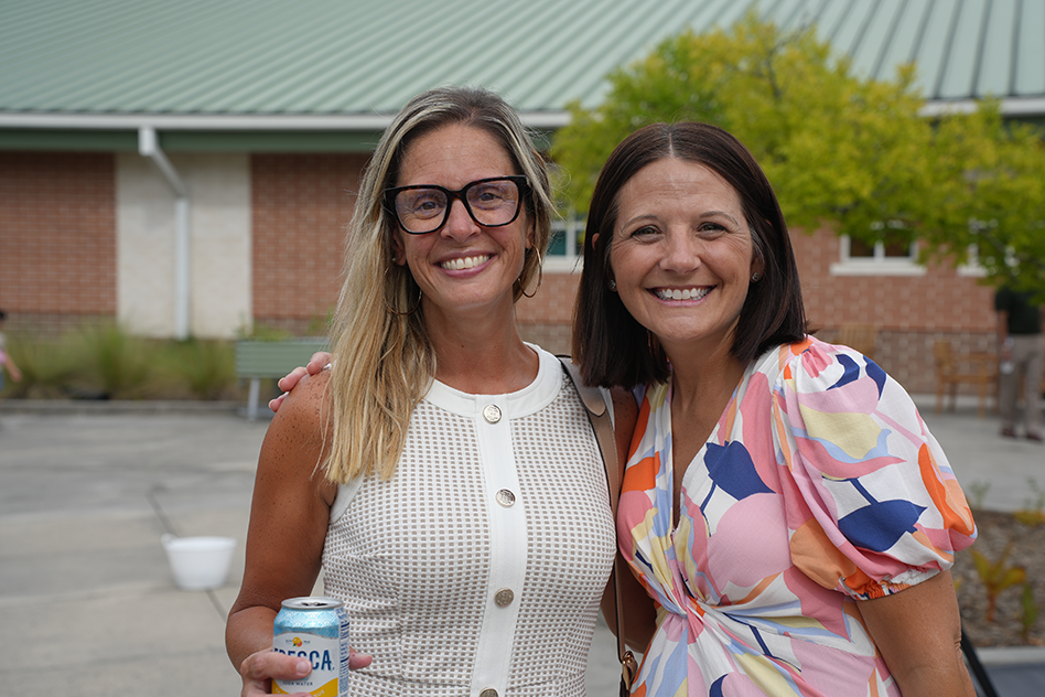 Two women smile in front of a brick building with a green roof. One holds a can.