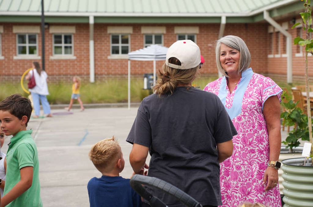 Adults and children stand in a parking lot. A woman in pink stands next to a woman in a cap.