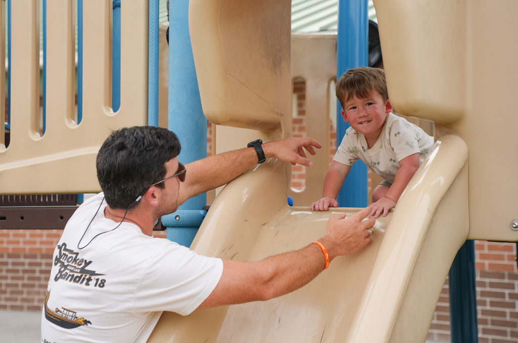 A man assists a young child in climbing up the slide in a playground. The slide is beige.