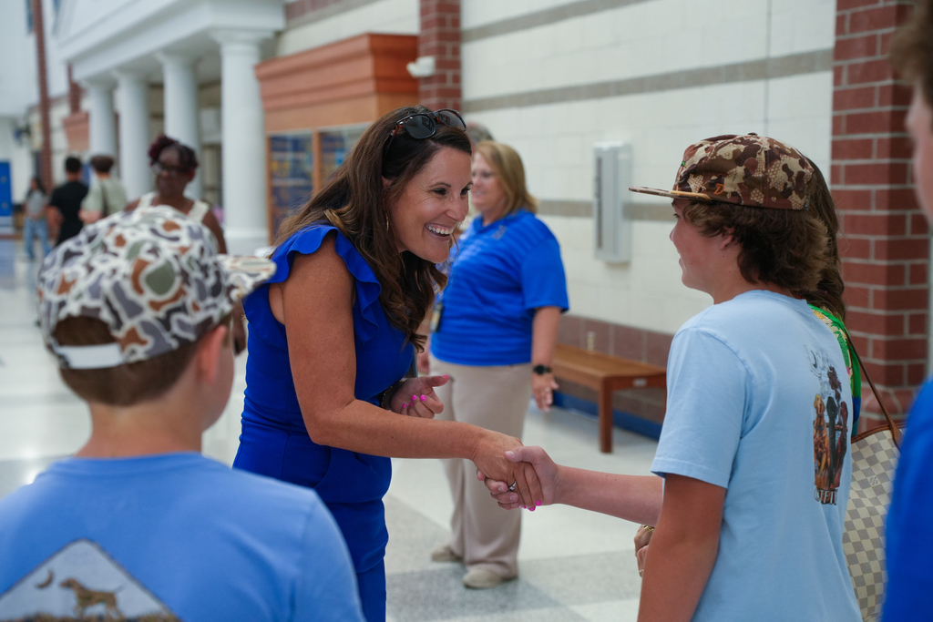People inside a hallway. A smiling woman in blue shakes hands with two young boys, one wearing a camouflage hat.