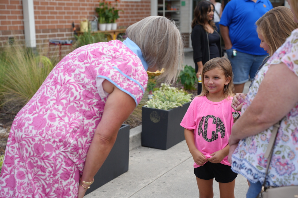 A woman in pink leans over a girl in a pink T-shirt. Behind them, another woman holds a handbag. In the background, people stand near a building with plants.