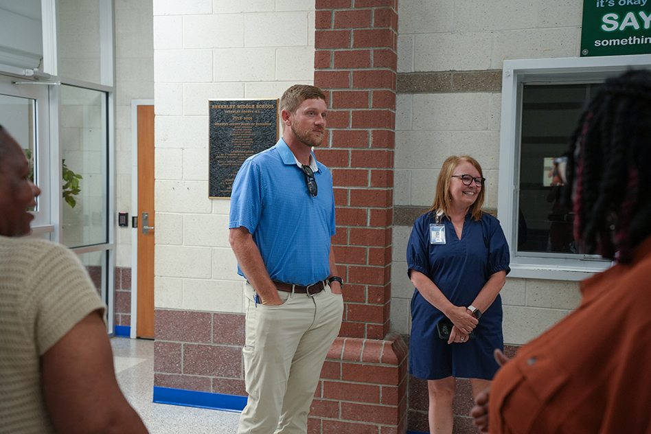 Four people stand in a corridor, with a brick wall and a sign behind them. A man wears a blue shirt, khaki pants, and a watch.