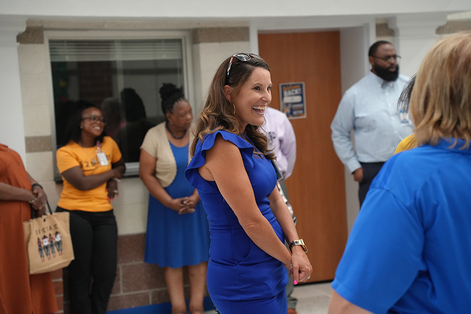 A woman in a blue dress stands and laughs in a hallway. Behind her, a group of people stands near a white wall and brown door.