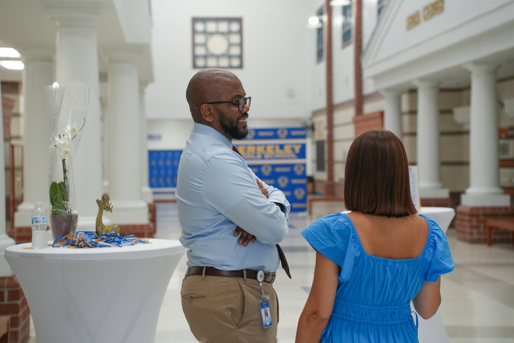 A man and a woman stand in an open hall, facing each other. The man wears glasses and a light blue shirt.