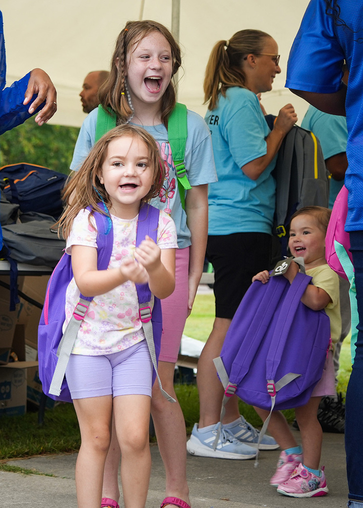 A group of children and adults with backpacks outdoors. One child wearing purple shorts smiles.