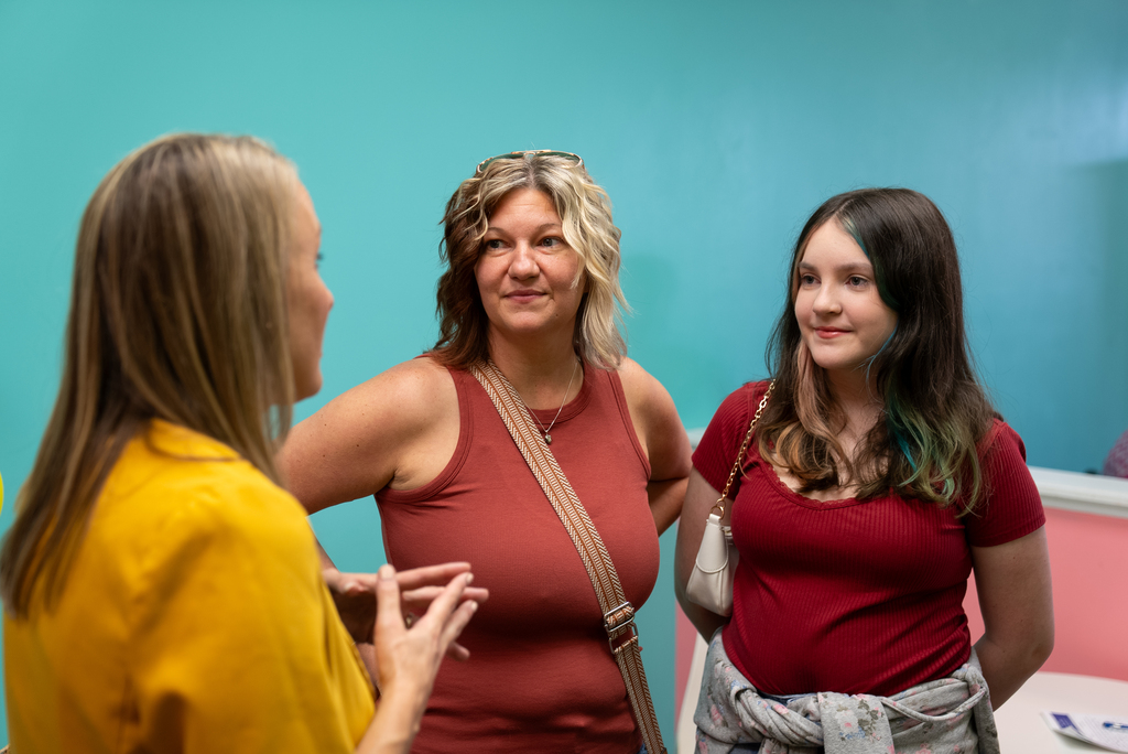 Three women in a room with a teal wall. They are facing each other, possibly engaged in a conversation.