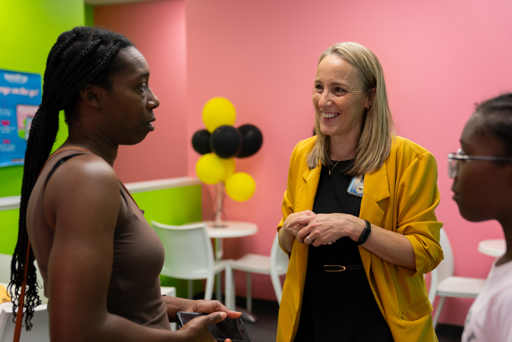 Three people converse in a colorful room with balloons, tables, and chairs. A woman with blonde hair smiles.