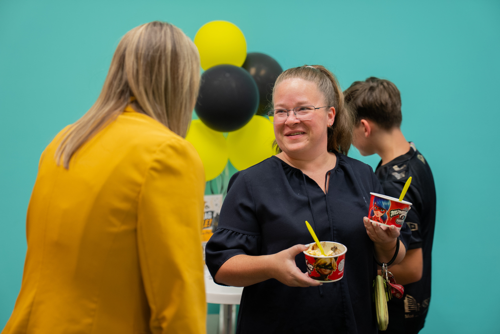 Three people stand before a turquoise wall with balloons; one woman holds a cup with a straw.