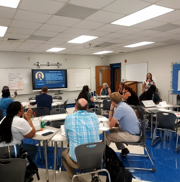 A classroom with blue flooring, tables, chairs, and a projector screen. Several people sit at tables, with a few standing.