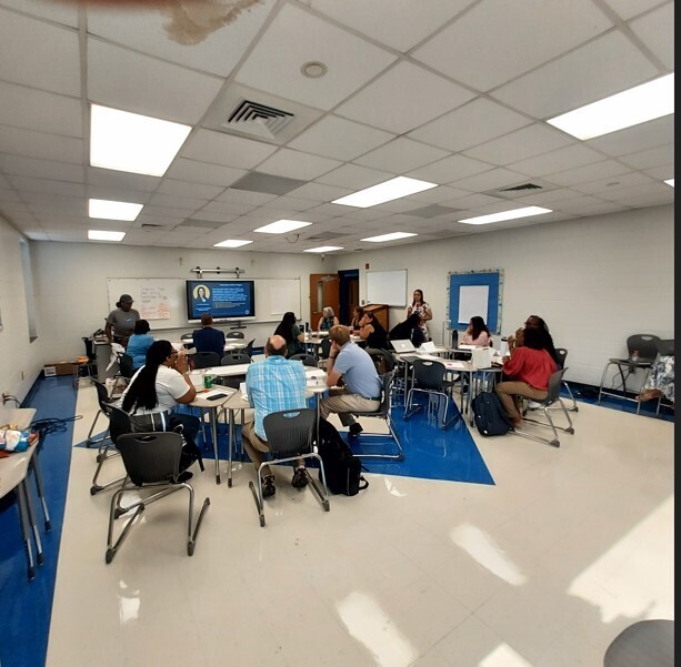 Group of people sitting around tables in a room with white walls and blue floor. A TV is on a stand.
