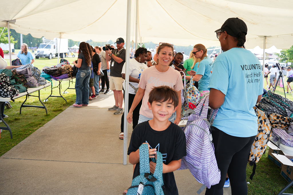 Several people gather under a white tent. A boy holds a backpack. A woman stands beside him.