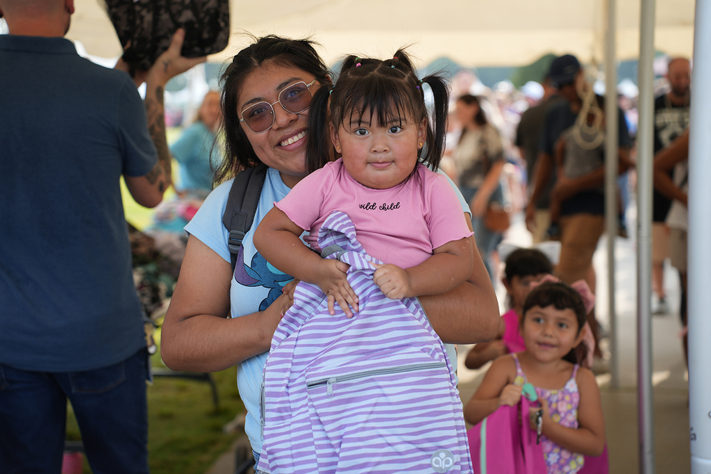 Woman holds a young girl and a backpack, smiling. A second young girl stands next to them.