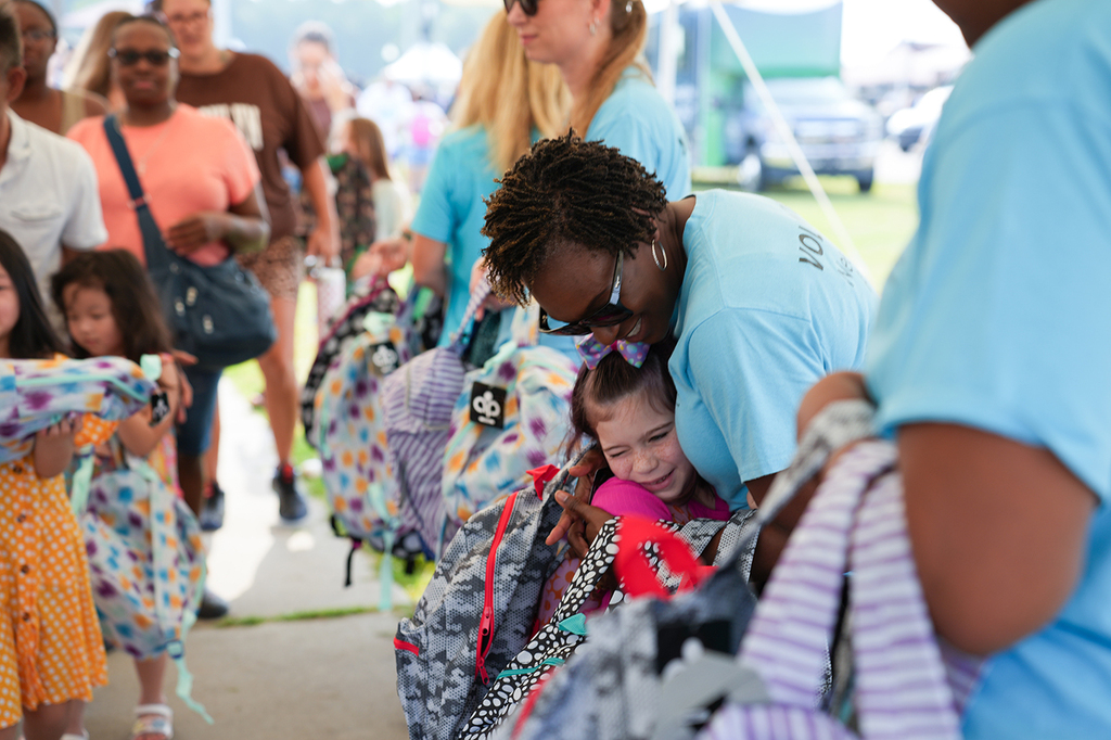 A crowd of people, some carrying bags, stands in an outdoor area. An adult helps a child with a backpack.