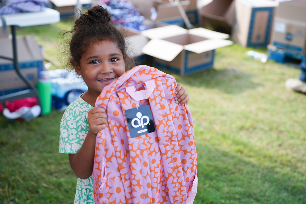 A child holds a pink backpack with an orange floral design, smiling, on a grassy field.