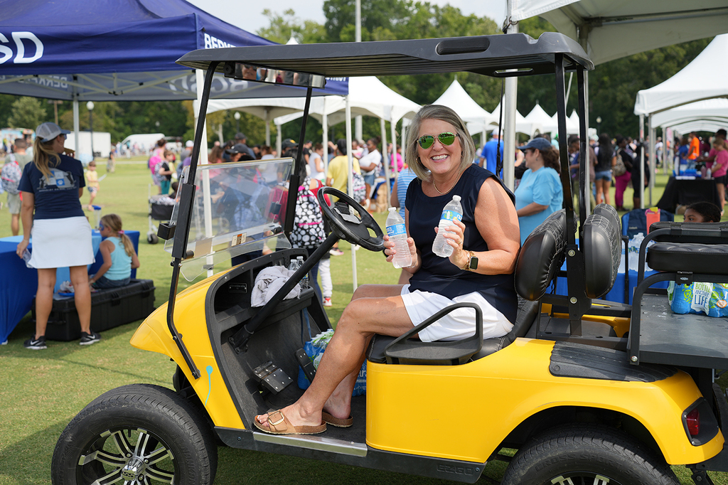 Woman in sunglasses and sandals seated in a yellow golf cart, with a bottle of water.