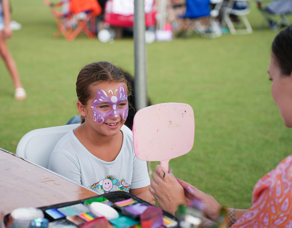 A girl with purple butterfly face paint holds a mirror to see her face. A woman with a bracelet helps.