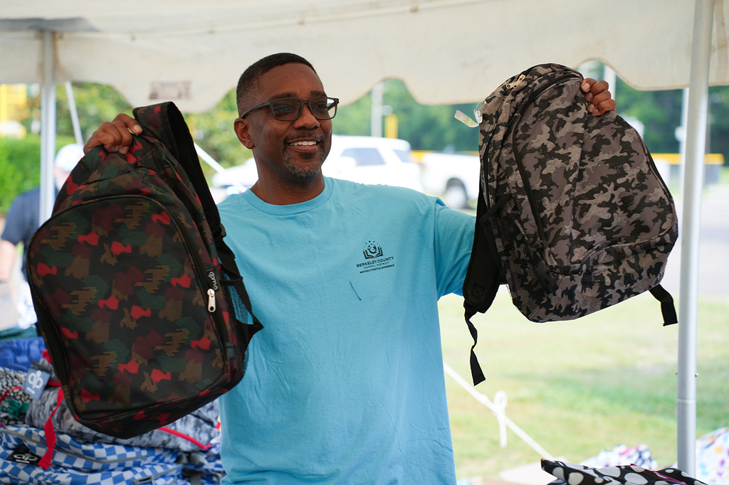 A man in a light blue t-shirt holds two camouflage backpacks up. A white tent is behind him.